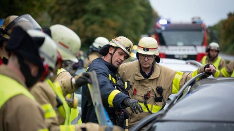 Mehrere Feuerwehrleute stehen neben einem Unfallauto und beraten sich. Im Hintergrund ist ein Feuerwehrauto zu sehen.